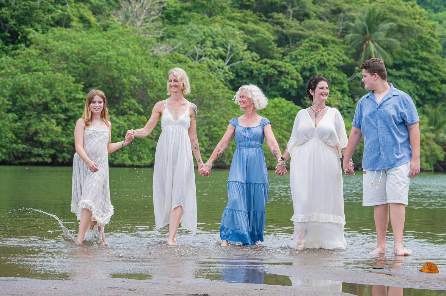 Five people holding hands wading in shallow water with lush green trees in the background.