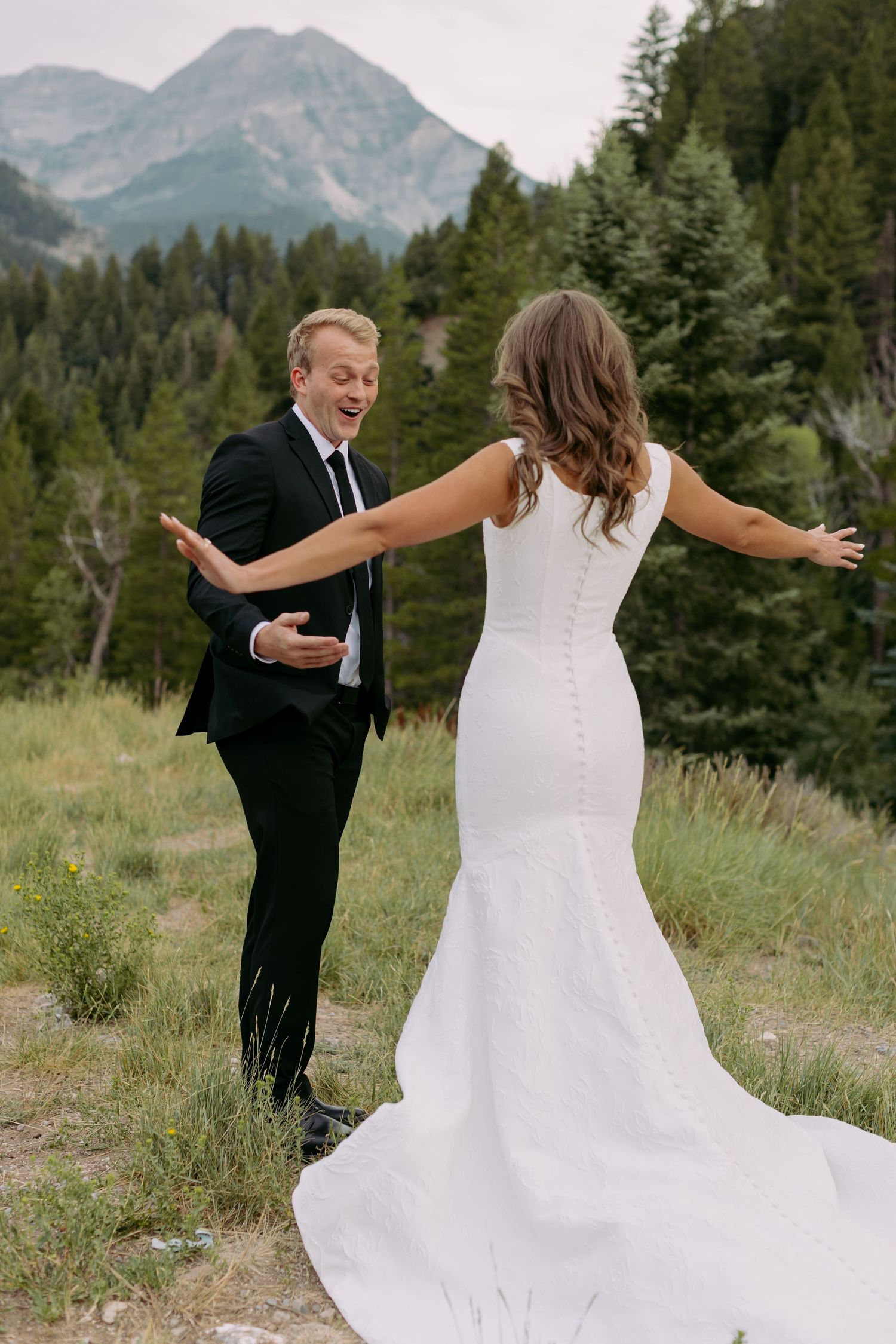 Groom in black suit reacts with joy seeing bride in white gown during first look with mountain backdrop.