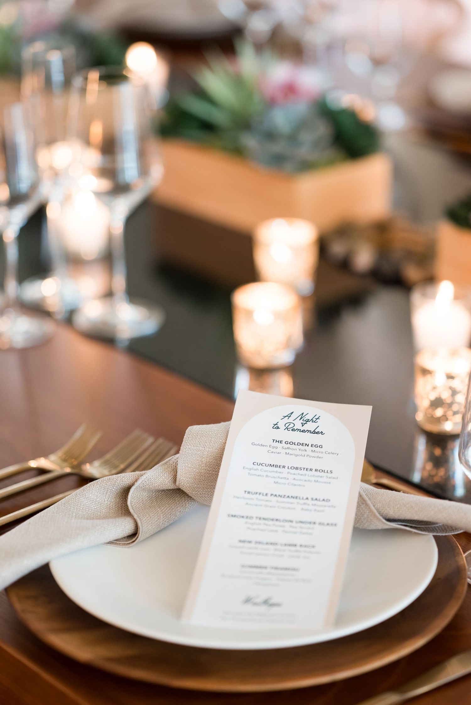 Elegant wedding table setting with menu card, linen napkin, white plate, gold cutlery, candles, and succulent centerpiece.