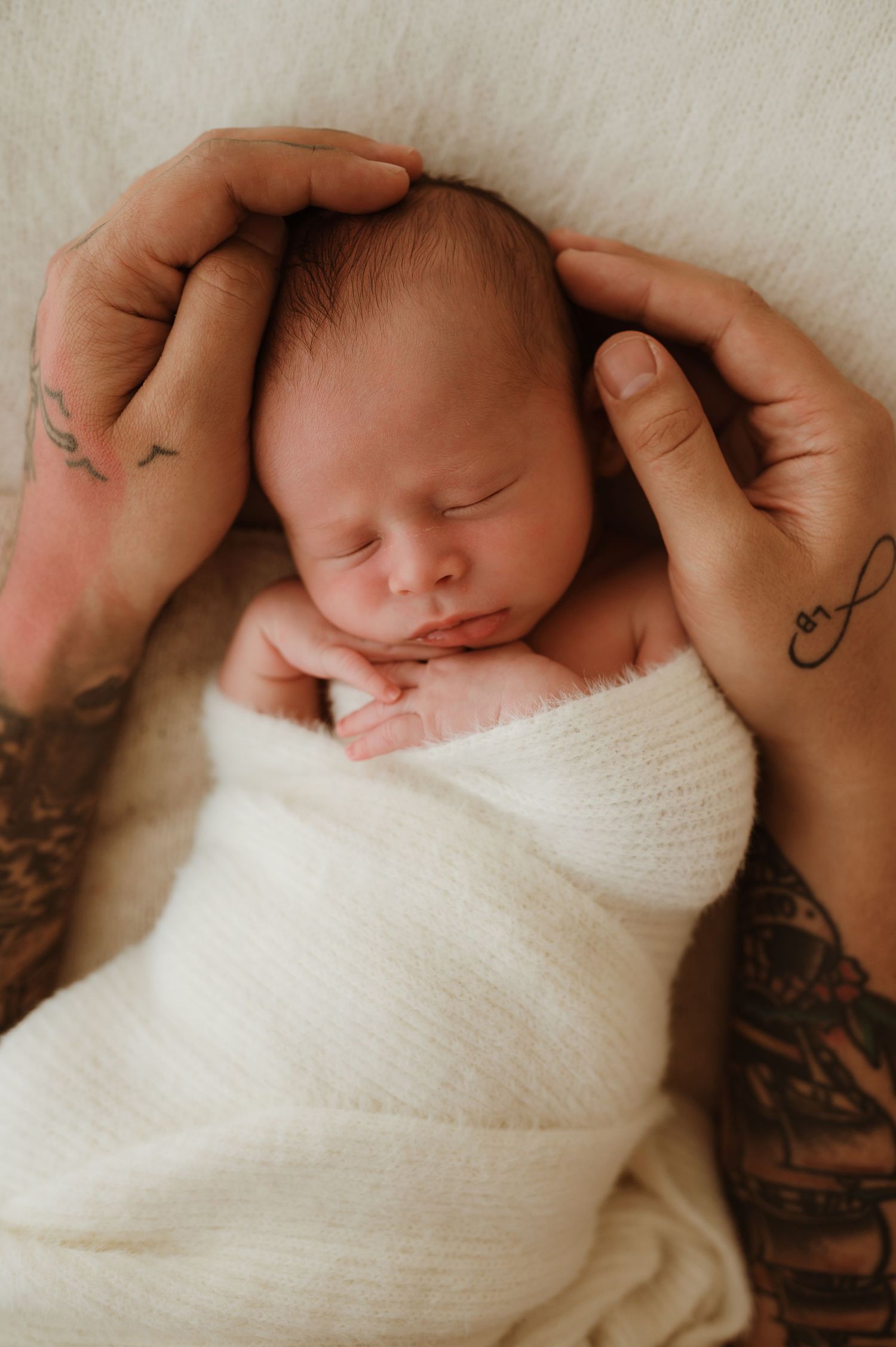Tattooed hands cradling a sleeping newborn baby wrapped in a soft white knit blanket.
