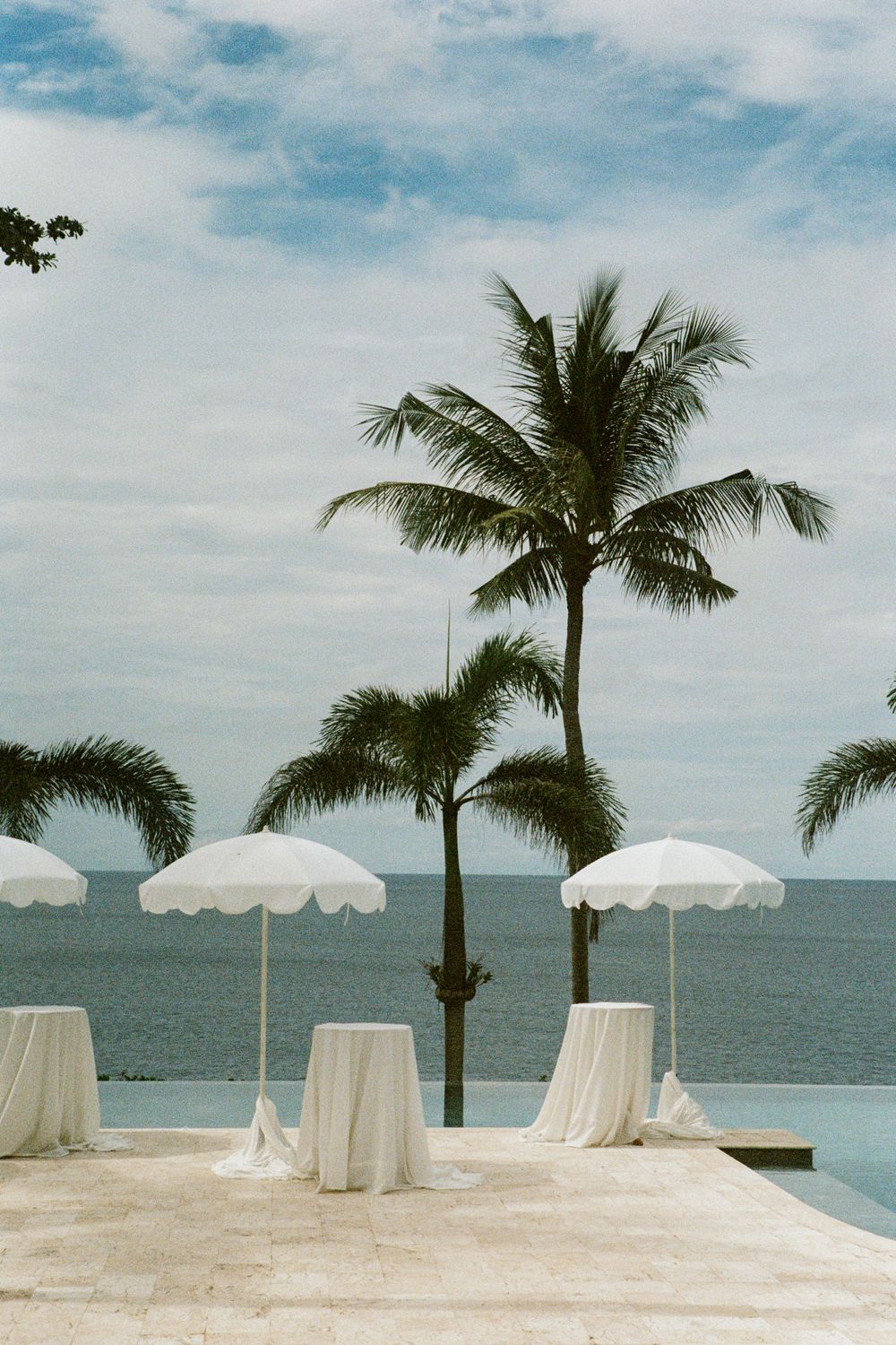 White umbrellas and cocktail tables line an oceanfront terrace with palm trees against a blue sky.