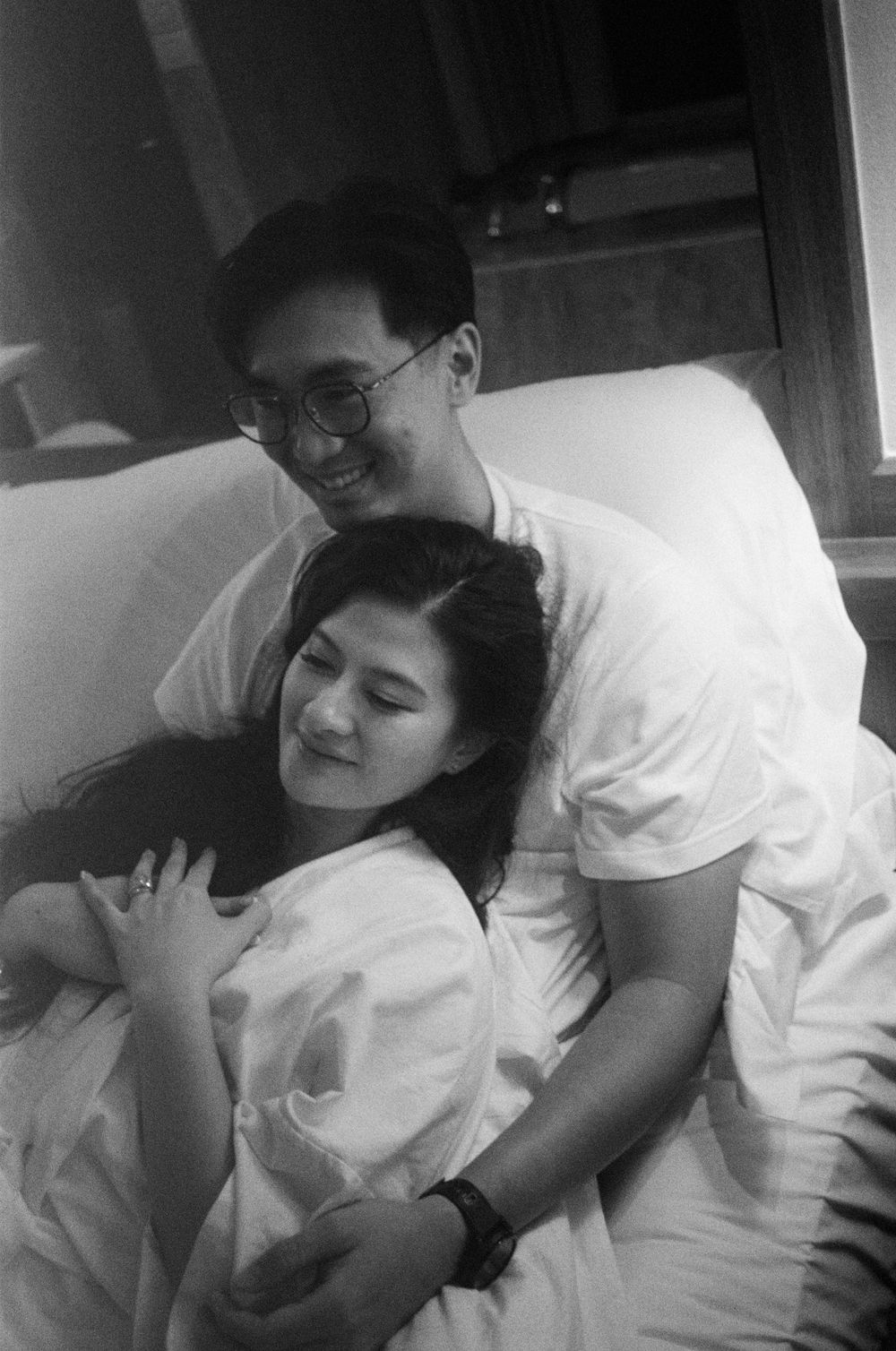 A tender black and white photograph of a couple embracing on white bedding.