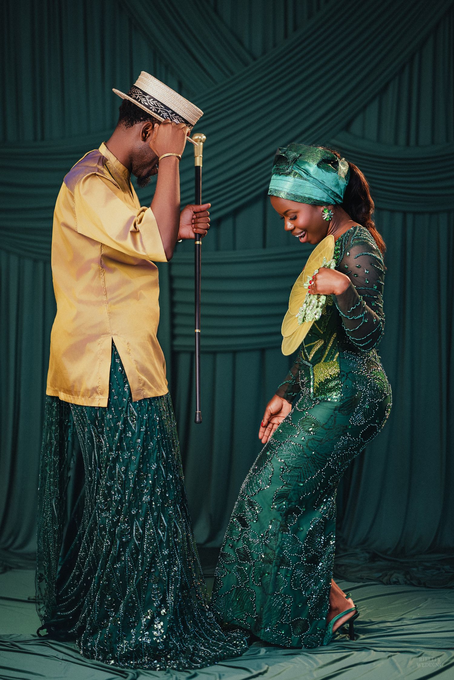 A couple in elegant green and gold African attire pose gracefully against a teal draped backdrop in a studio setting.