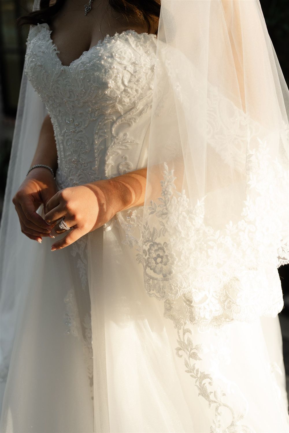 Bride in elegant white lace wedding gown with veil, showcasing intricate floral embroidery and a sparkling ring.