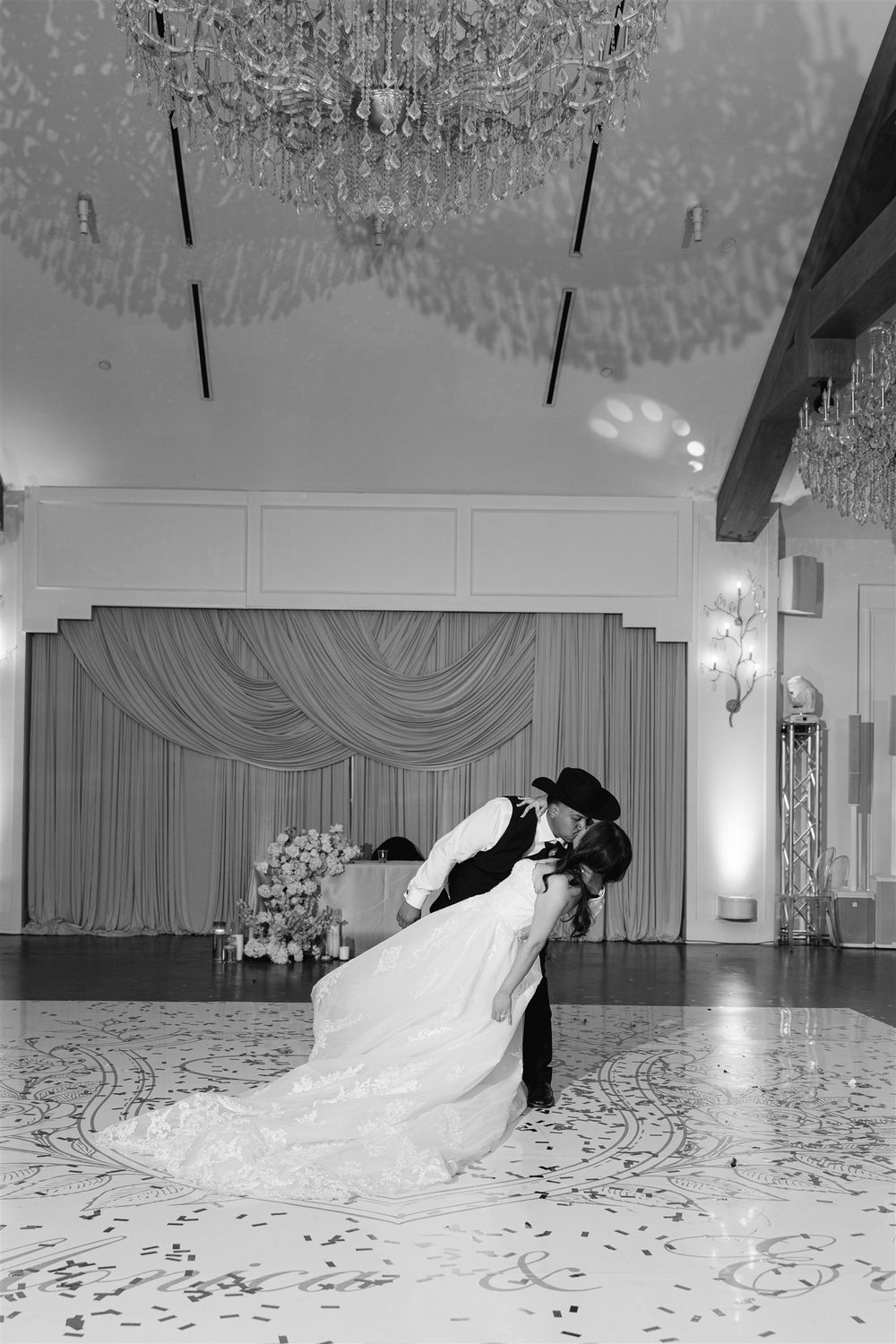 Bride and groom share romantic first dance dip in elegant ballroom with crystal chandeliers, black and white wedding photo.