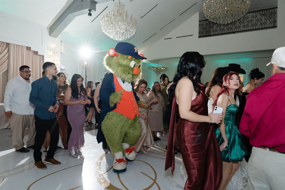 A green alligator mascot in a hat dances with elegantly dressed guests at a lively formal party event.