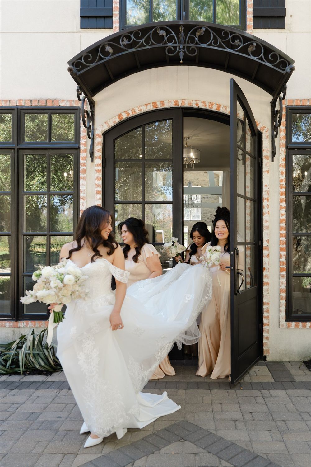 Bride in lace gown exits elegant brick venue doorway as bridesmaids in champagne dresses help carry her train.