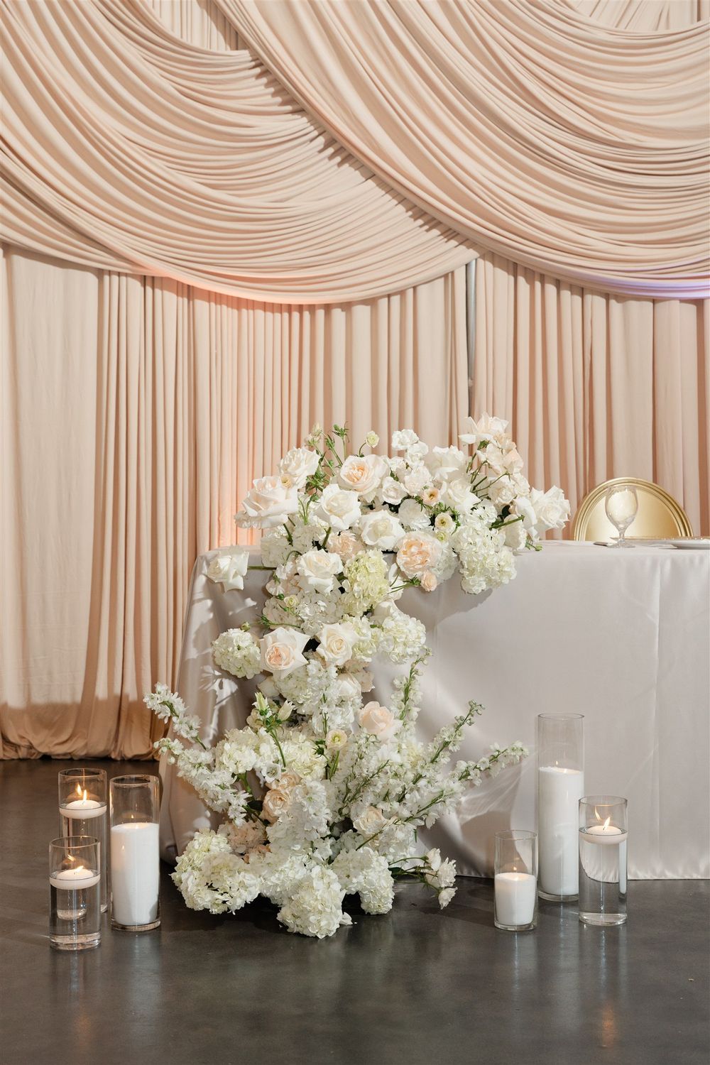 Elegant wedding sweetheart table with white floral arrangement, candles, and blush draped backdrop.