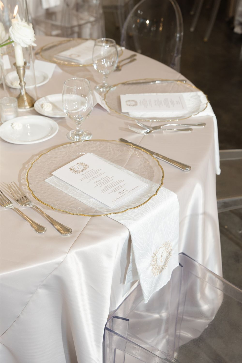 Elegant wedding reception table setting with gold-rimmed charger plates, crystal glassware, and white linen tablecloth.