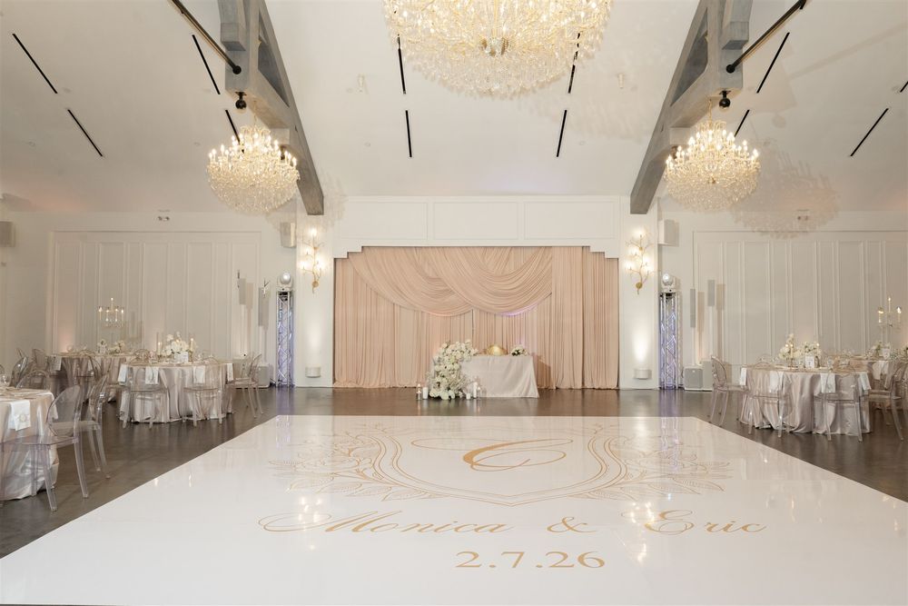 Elegant wedding ballroom with white dance floor monogram, chandelier lighting, draped backdrop, and reception tables.