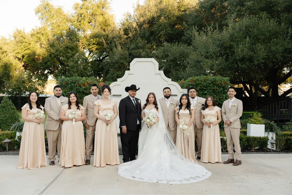 Wedding party poses outdoors in neutral beige attire with bride in white gown and groom in black cowboy hat.
