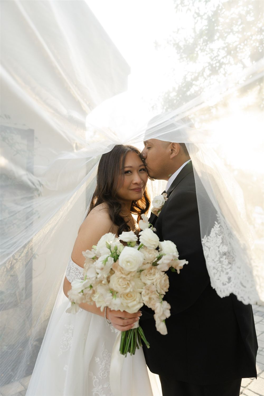 Bride and groom share a tender kiss under a flowing white veil, bride holding a lush white peony bouquet.