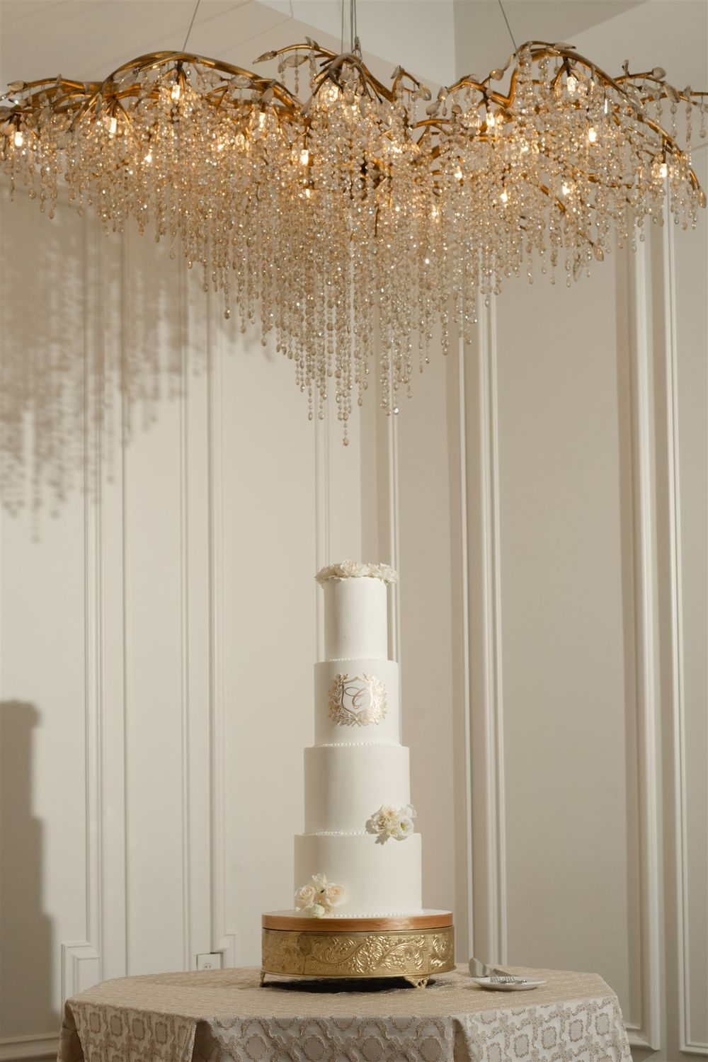 Elegant five-tier white wedding cake with gold details displayed under a crystal chandelier.