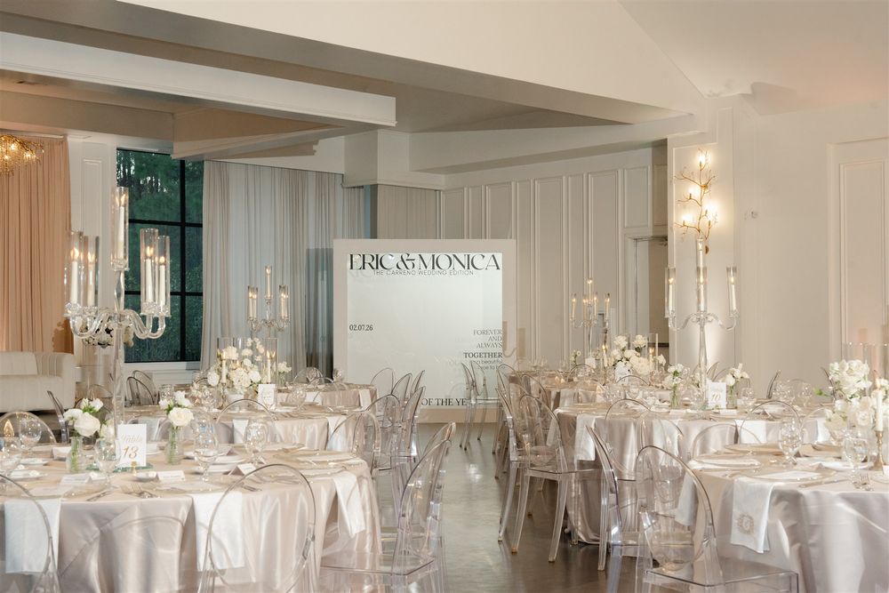 Elegant white wedding reception hall with ghost chairs, floral centerpieces, candelabras, and a personalized backdrop sign.