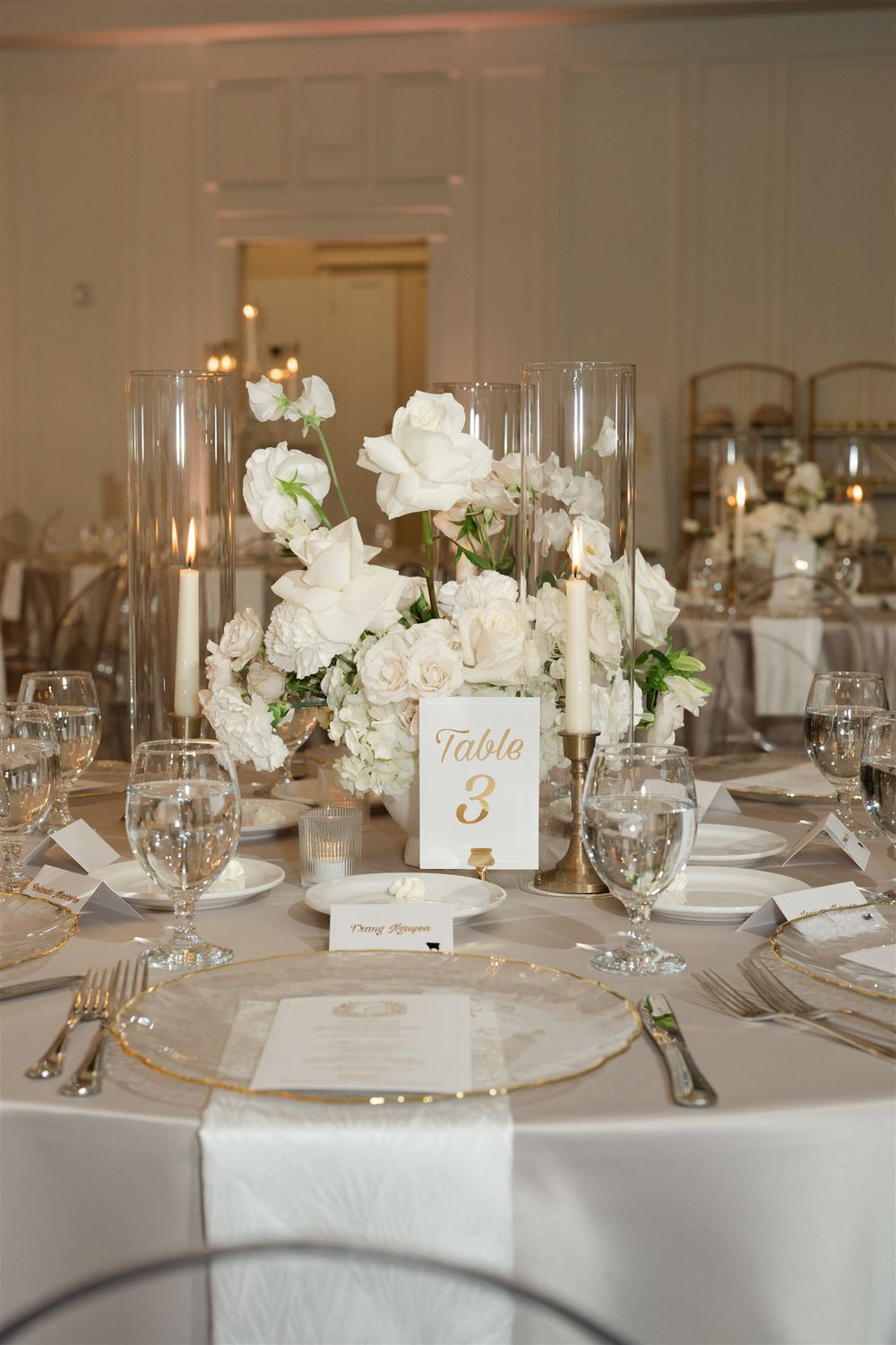Elegant wedding reception table with white floral centerpiece, tall candles, gold charger plates, and Table 3 sign.