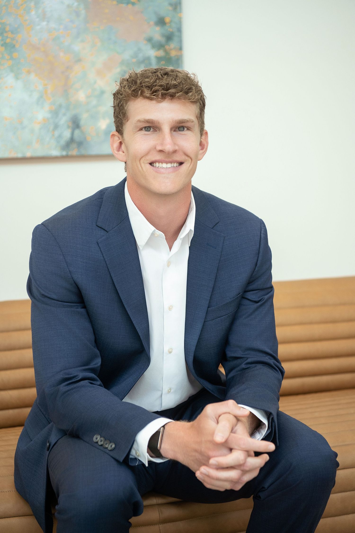 Headshot of young businessman in blue suit sitting relaxed with hands clasped in modern office setting.