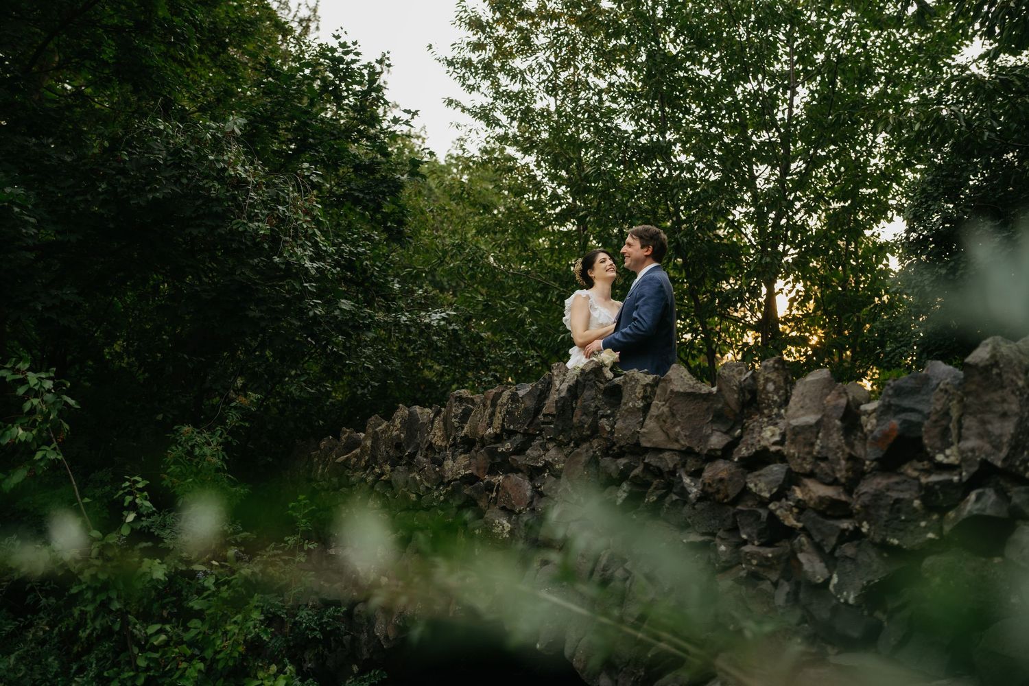 A couple in wedding attire sits on a rustic stone wall surrounded by lush green trees at golden hour.