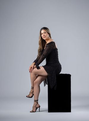 Elegant woman sitting on black block wearing black lace dress and strappy heels.