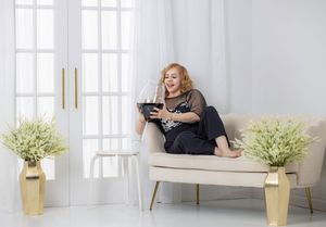 Cheerful older woman relaxing on beige sofa holding tablet in bright white room.