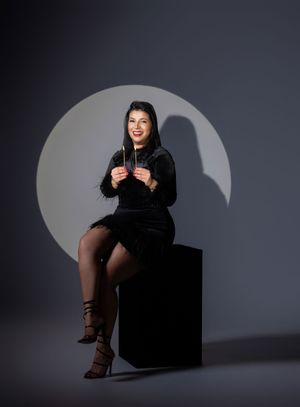 Woman in black velvet dress sitting on cube pedestal, smiling, holding sparklers against gray studio backdrop.