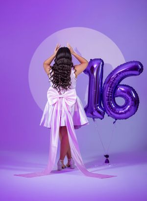 Girl in white satin dress with bow poses back to camera near purple number 16 balloons.