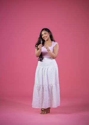 Young woman in white eyelet maxi dress smiles against pink background.