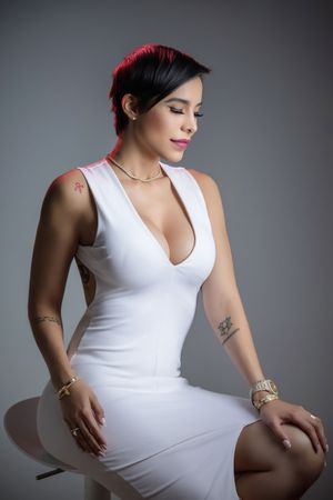Woman in white deep-V dress with tattoos and gold jewelry sits posed against gray background.