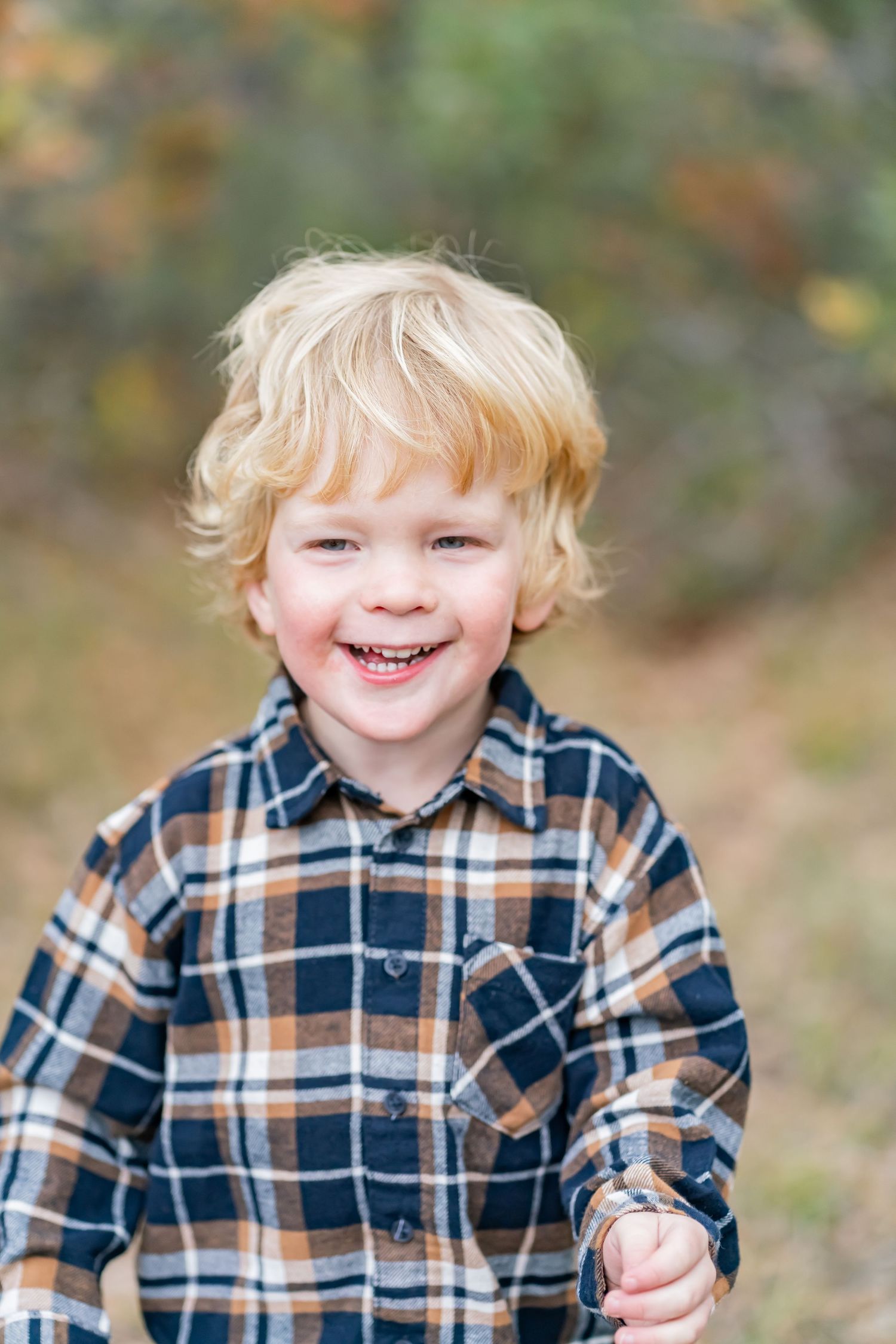 Smiling blond toddler boy in plaid flannel shirt laughing outdoors in autumn setting.