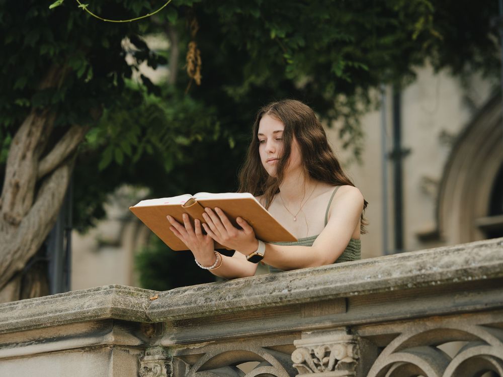 Young woman with long brown hair reading a book on a stone balcony surrounded by lush green trees.