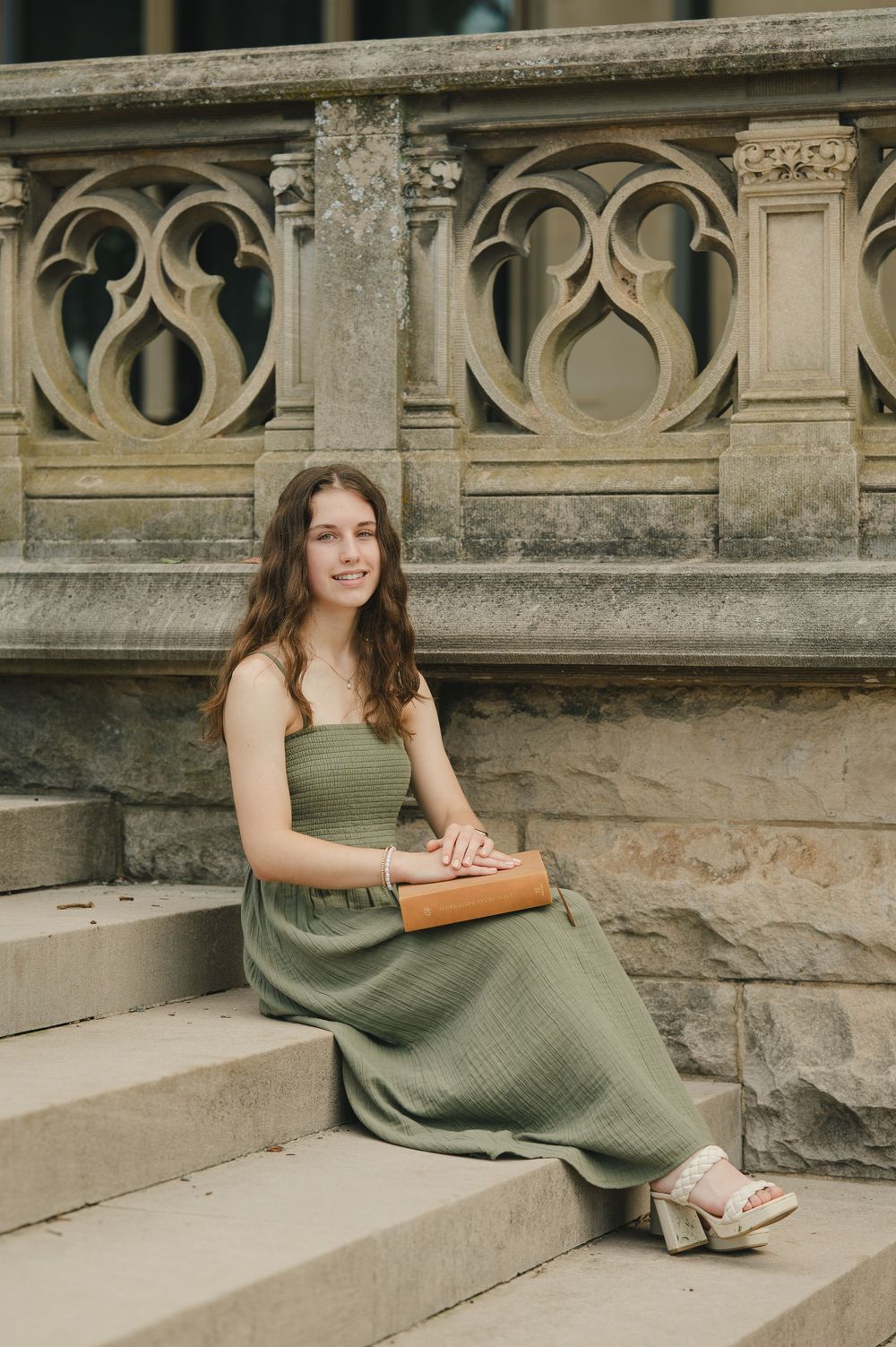 Young woman in green maxi dress sitting on stone steps holding a brown clutch, smiling outdoors.