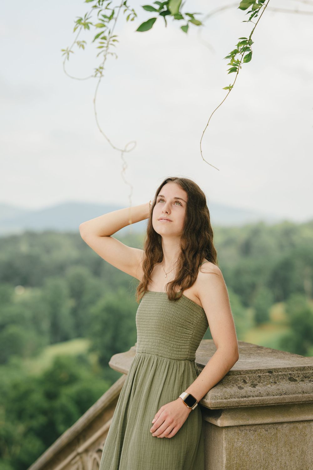 Young woman in olive green dress poses on stone balcony with lush green mountain landscape behind her.