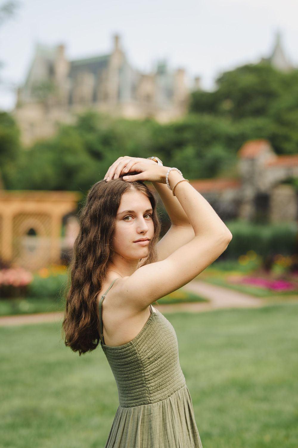 Young woman in olive green dress poses gracefully in lush garden with historic mansion in background.