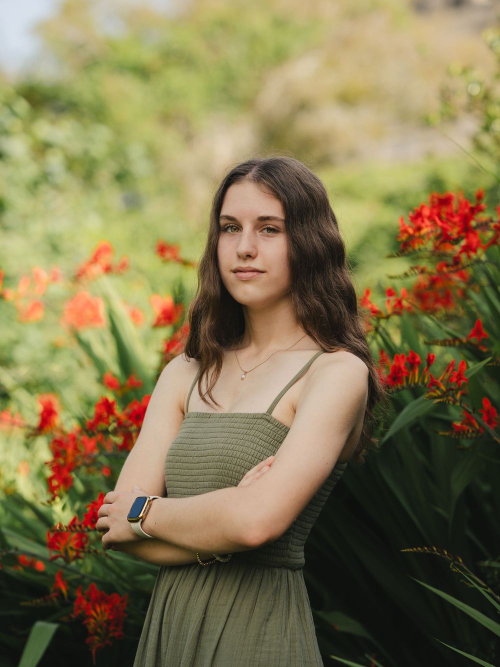 Young woman in olive dress with arms crossed standing among vibrant red flowers in a lush garden.