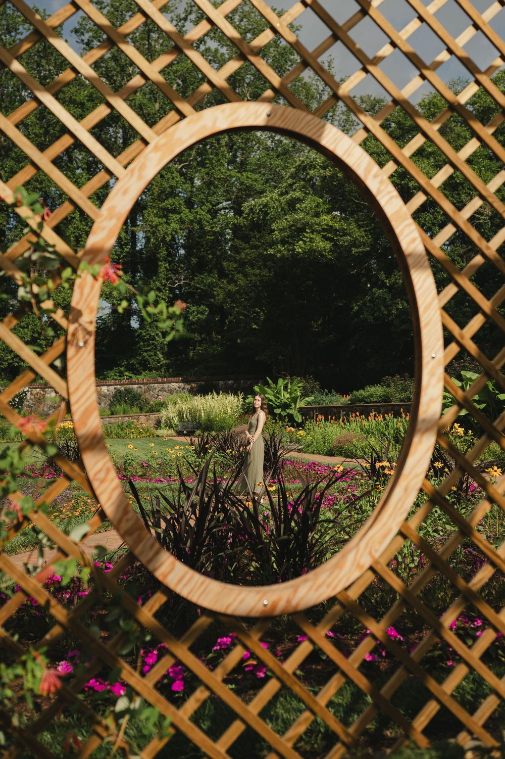 Person reflected in oval mirror mounted on wooden lattice trellis surrounded by colorful garden flowers and green trees.