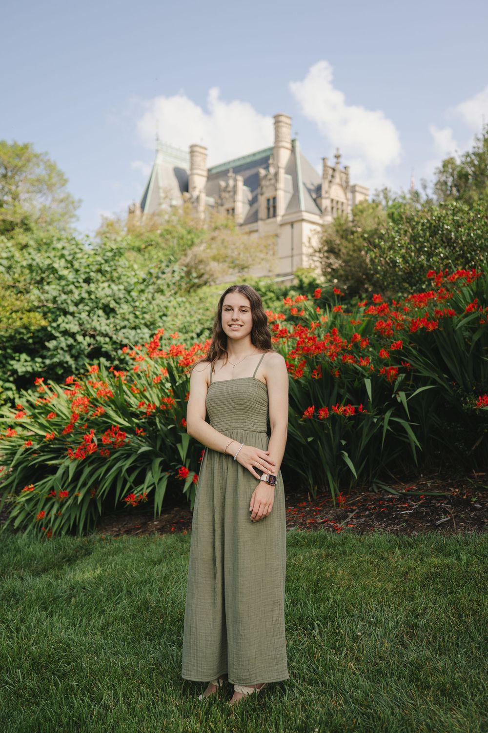 Young woman in olive green jumpsuit poses in front of red flowers and a grand estate building on a sunny day.