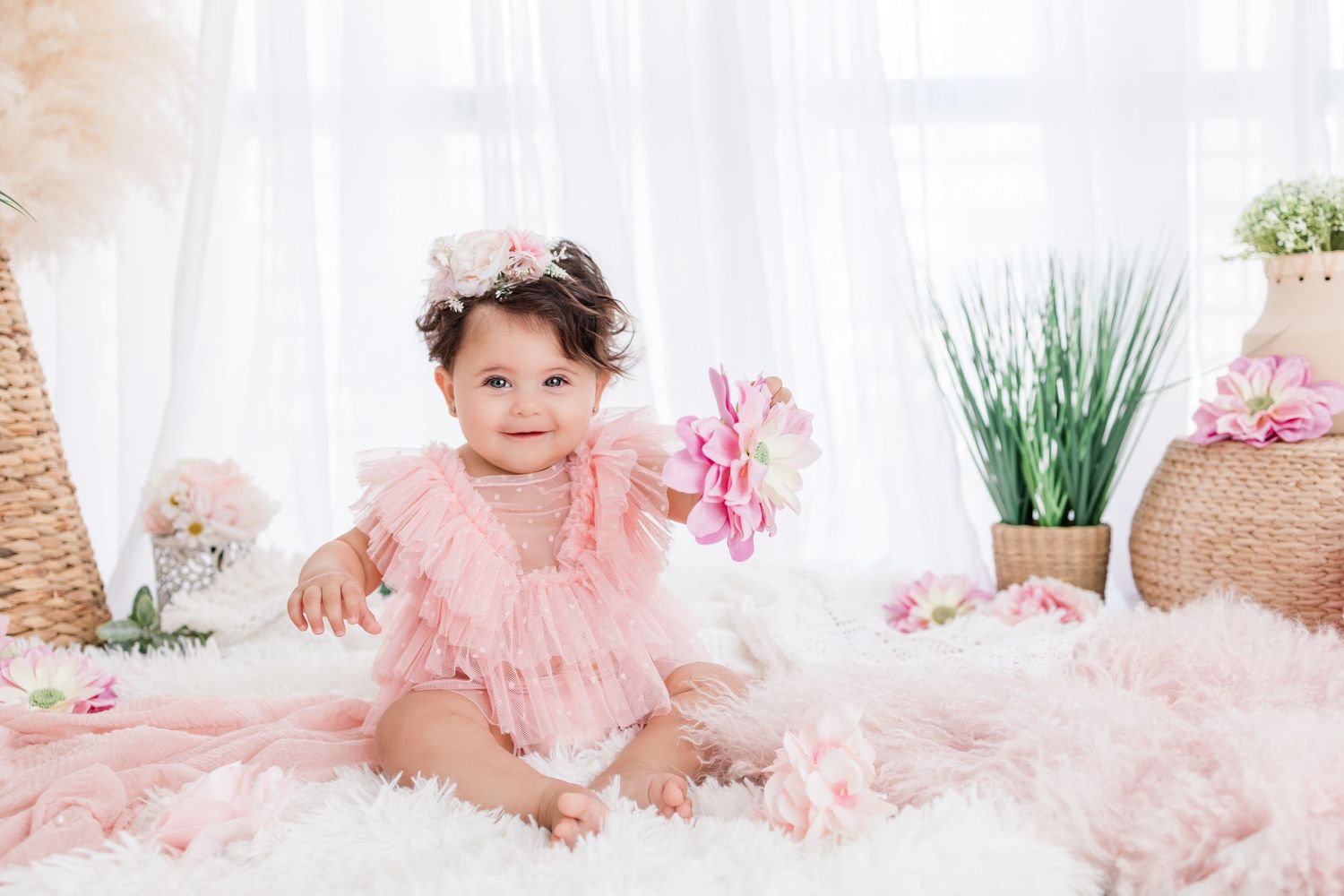 Adorable baby girl in lace romper and floral headband lying on blanket, smiling during professional photo session.