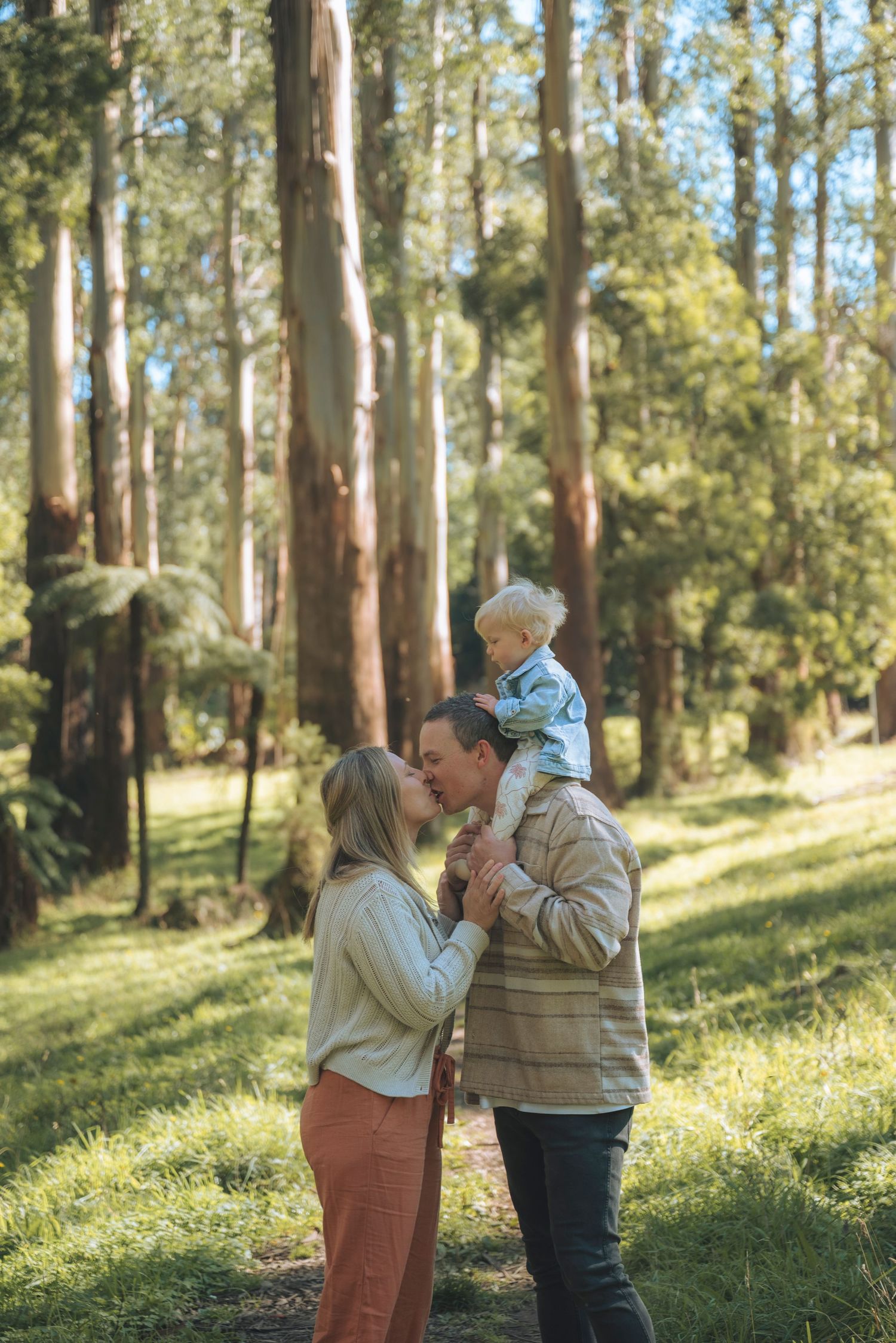 A couple kisses while holding their toddler in a sunlit forest clearing with tall eucalyptus trees.