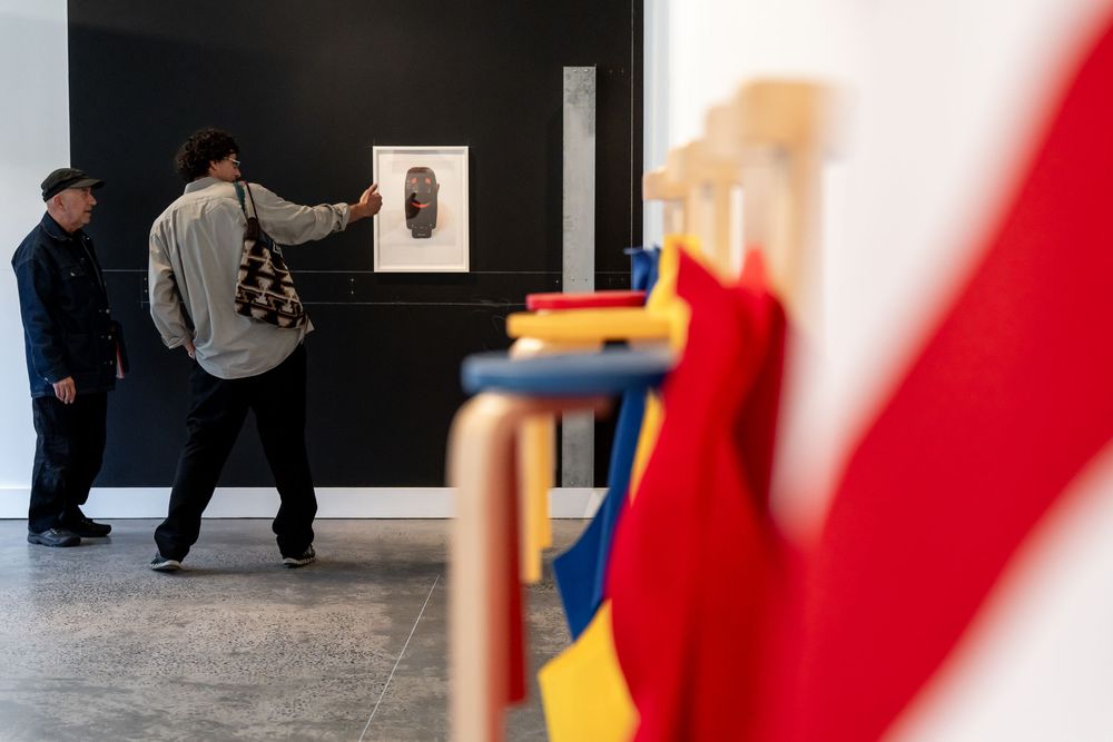 Two visitors examine artwork on a gallery wall while colorful flags hang in the foreground of a modern art exhibition.