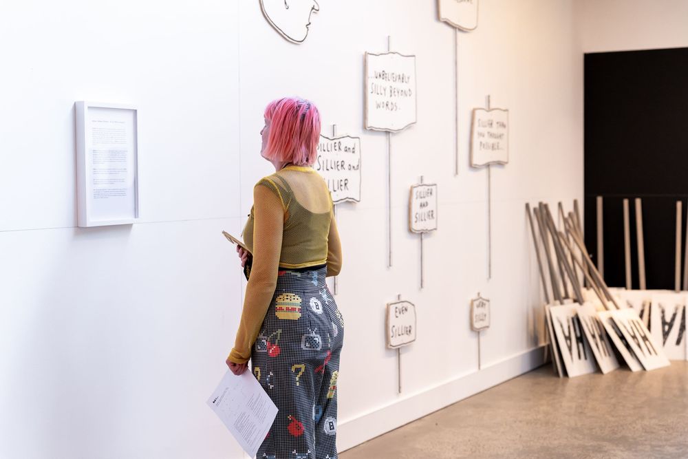 Woman with pink hair viewing handwritten notes on gallery wall beside wooden frames in modern art exhibition.