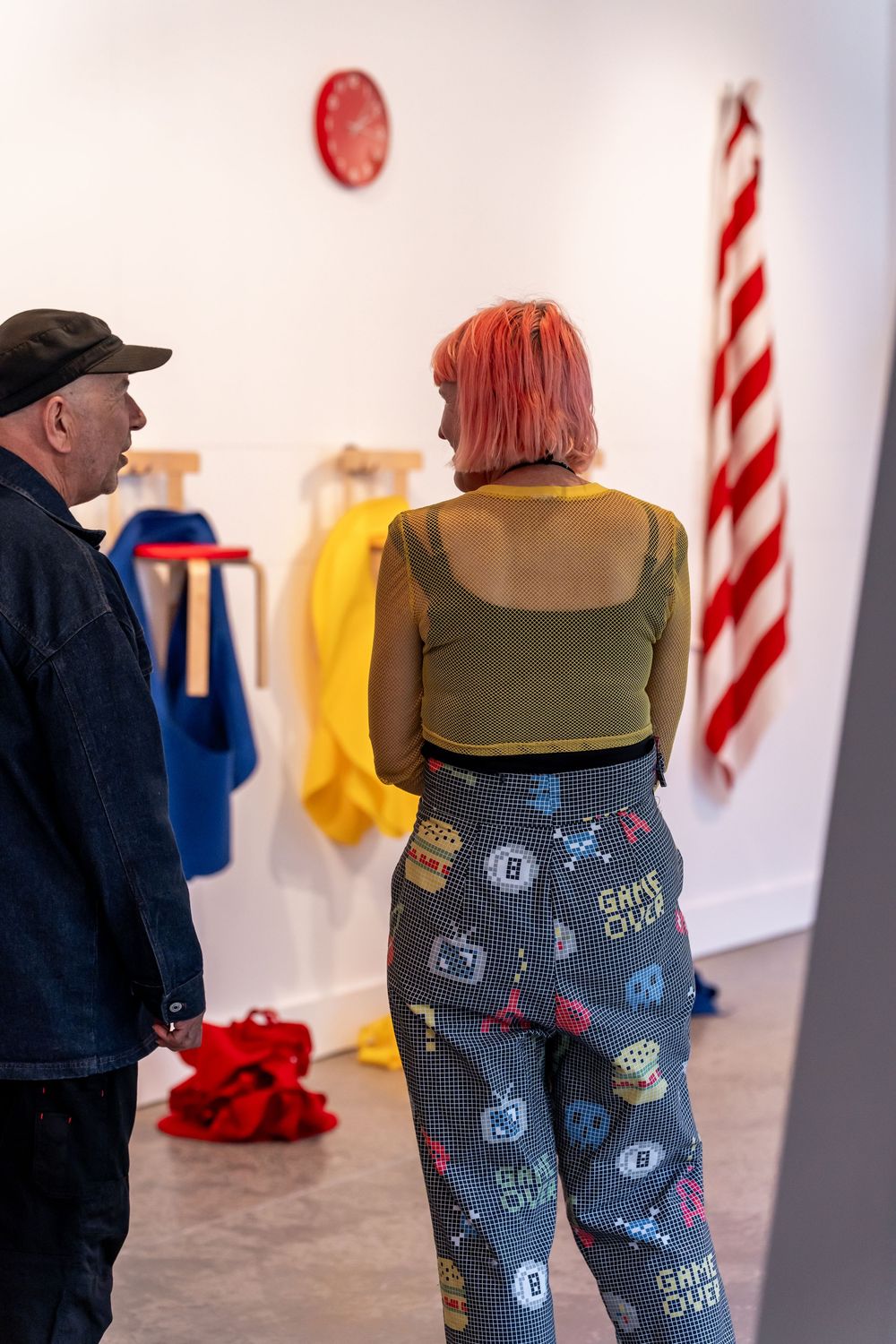 Two people viewing colorful art installations in a gallery, woman with pink hair wearing game-over patterned pants.