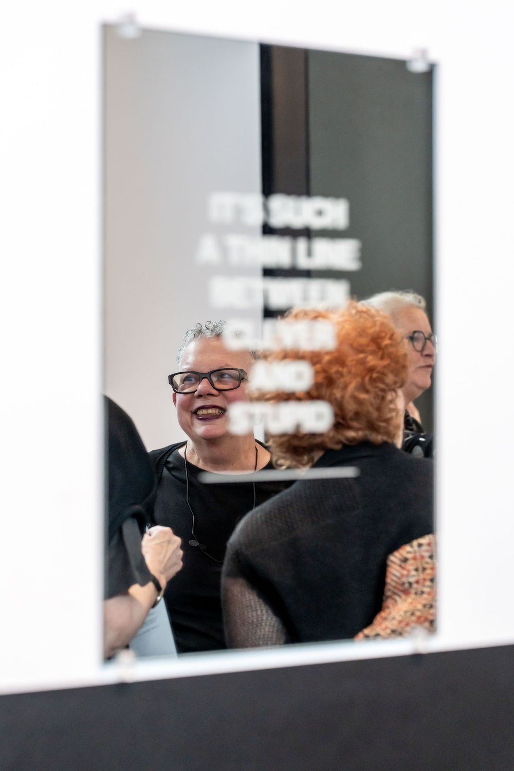 Two women laugh together reflected in a mirror with bold text at an art gallery exhibition.