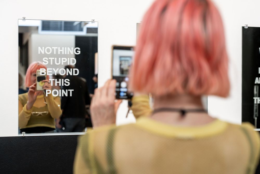 Person with pink hair photographing a mirror sign reading 'Nothing Stupid Beyond This Point' at an art gallery.