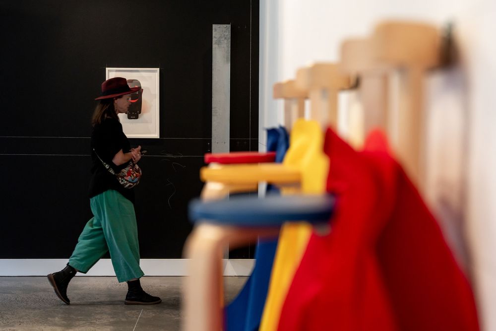 Woman in green pants and hat walks past colorful coats hanging on wooden hooks in a modern gallery space.