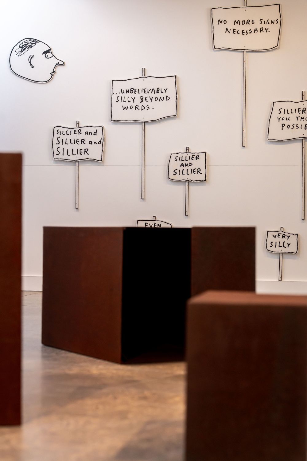 Art installation with protest signs reading silly phrases on white wall, brown metal pedestals in foreground, gallery space.
