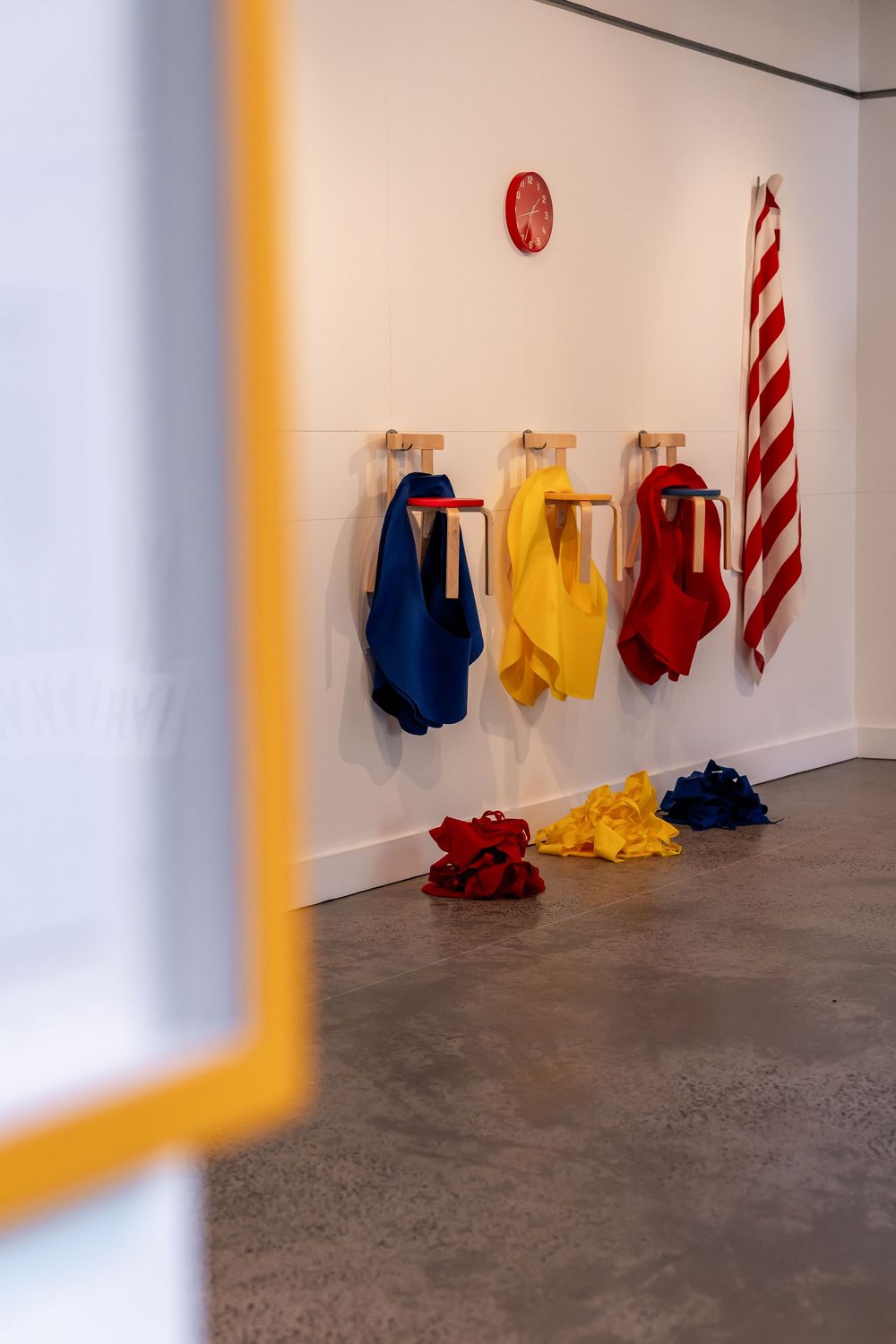 Colorful blue, yellow, and red garments hang on wooden pegs in a gallery, with matching fabric piles on the floor.
