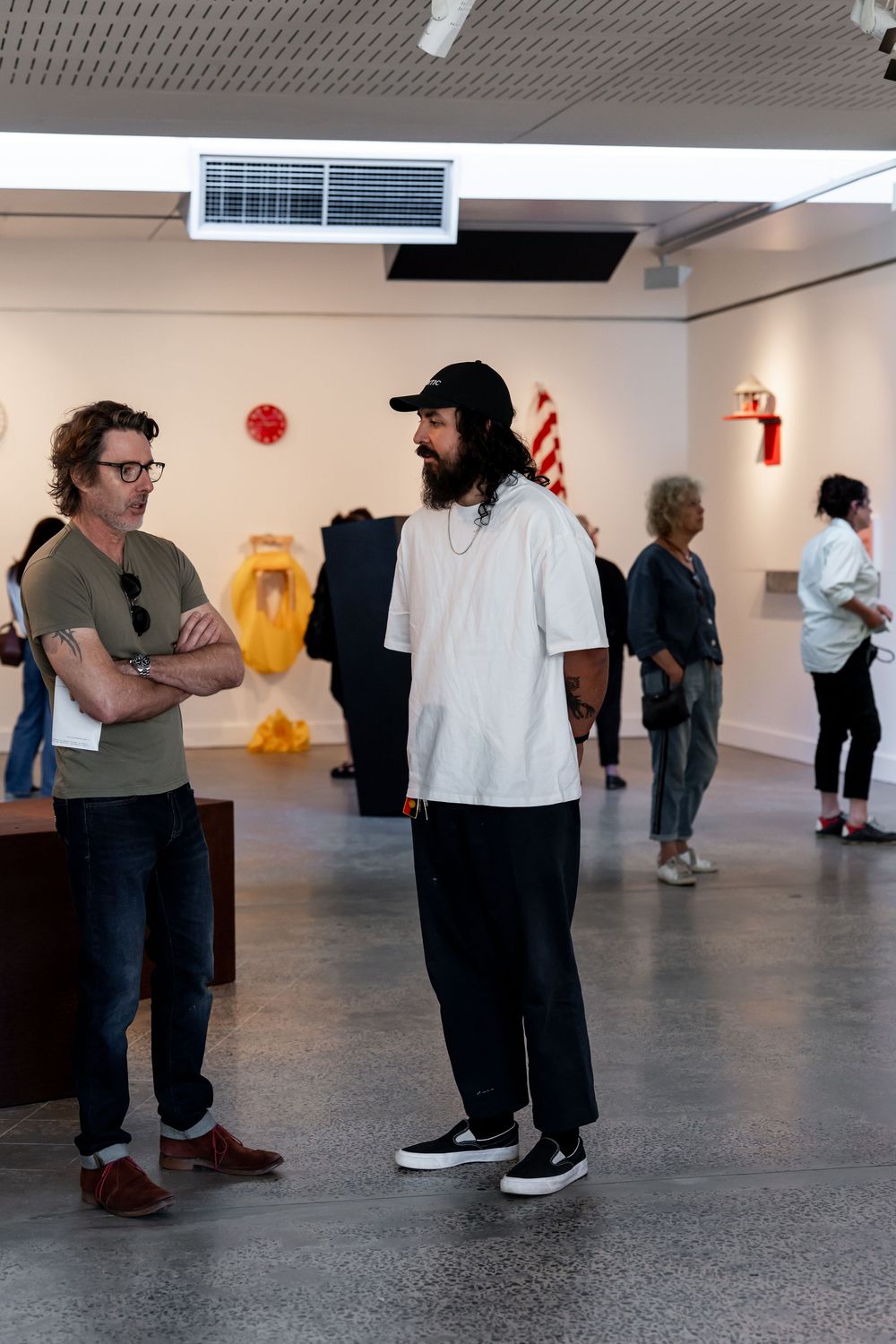 Two men conversing in a modern art gallery surrounded by colorful sculptures and other visitors.
