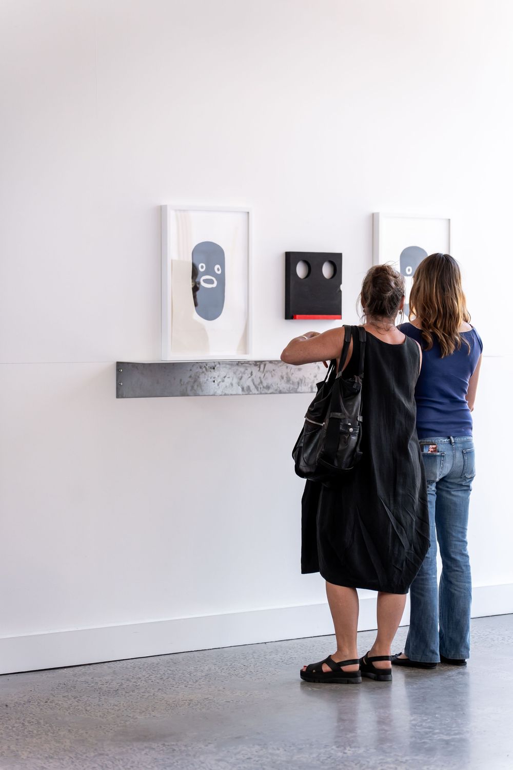 Two women viewing abstract face artwork on a white gallery wall with a metal shelf display.