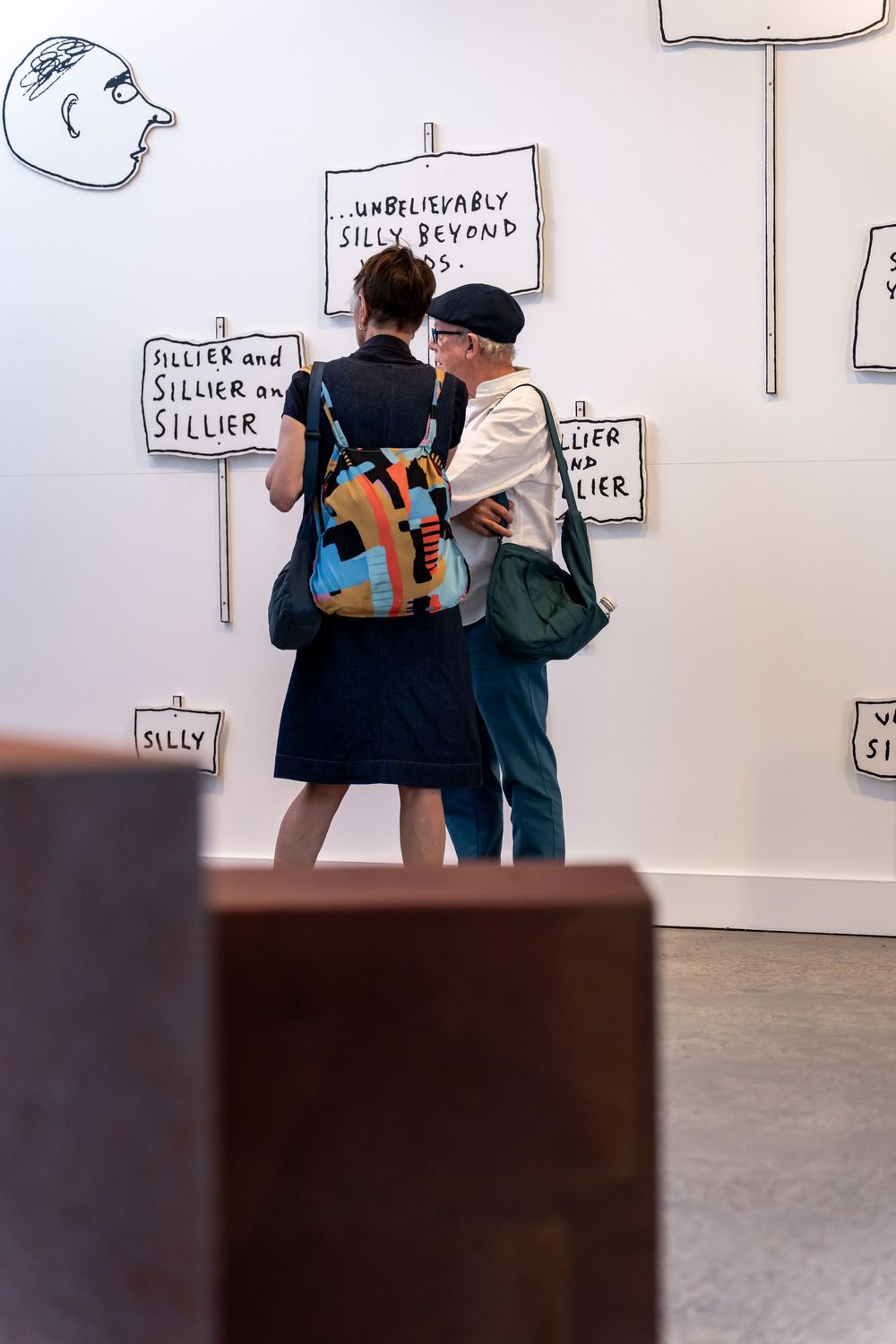 Two museum visitors with bags examine silly-themed text art installations on a white gallery wall.