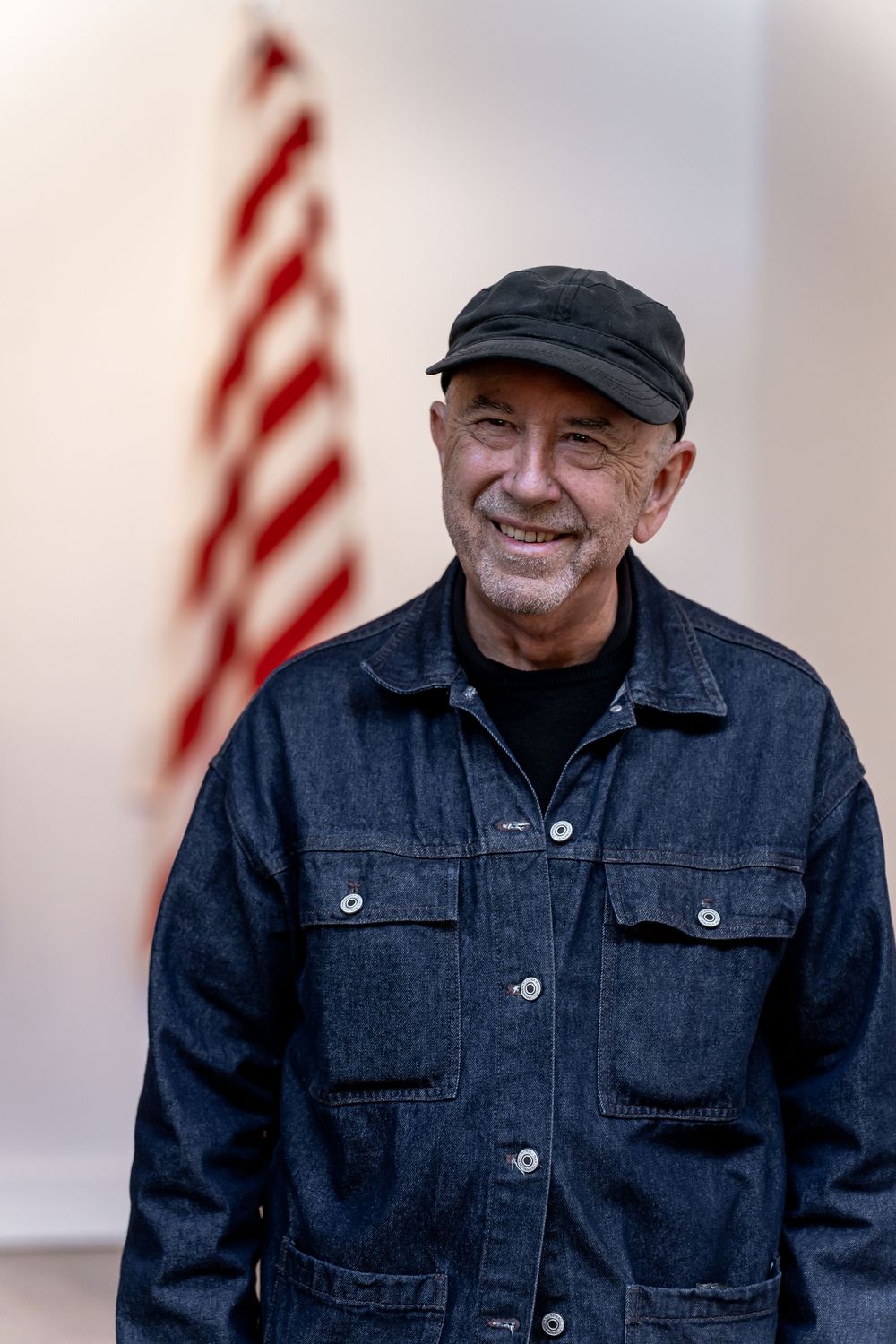 Smiling senior man in denim jacket and cap standing before an American flag backdrop.