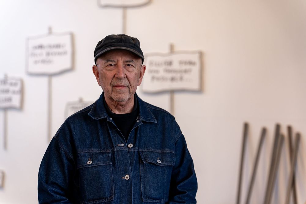 Elderly man in denim jacket and black cap stands before white wall with hanging signs in art gallery.